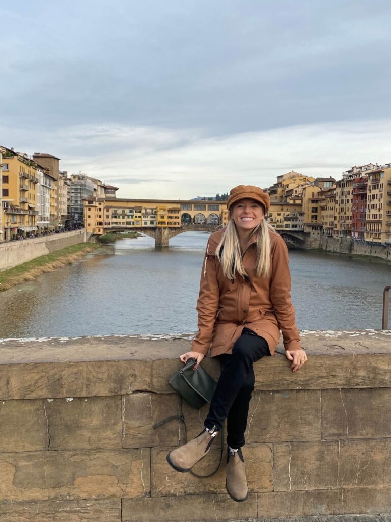 Chelsey Explores sitting on a bridge smiling with the Ponte Veccio bridge in the background